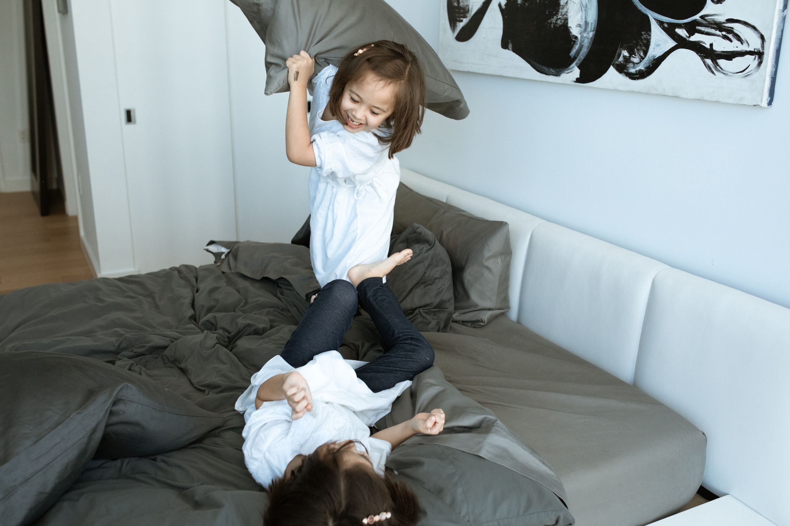 Two young sisters enjoy a fun pillow fight in a cozy modern bedroom setting.
