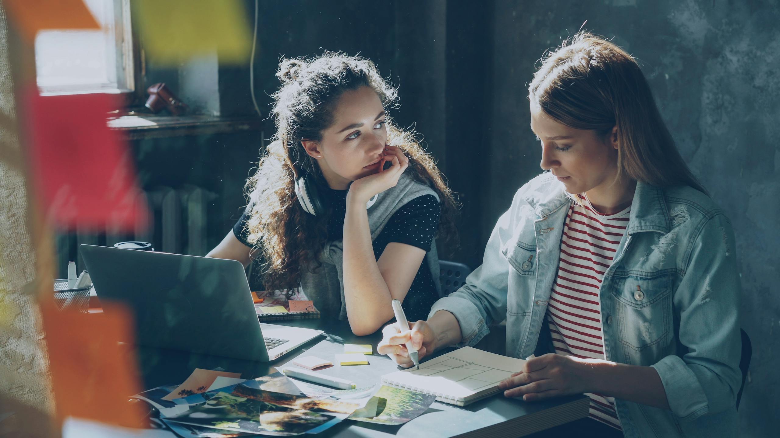 Two young women collaborating and brainstorming at a desk with a laptop.