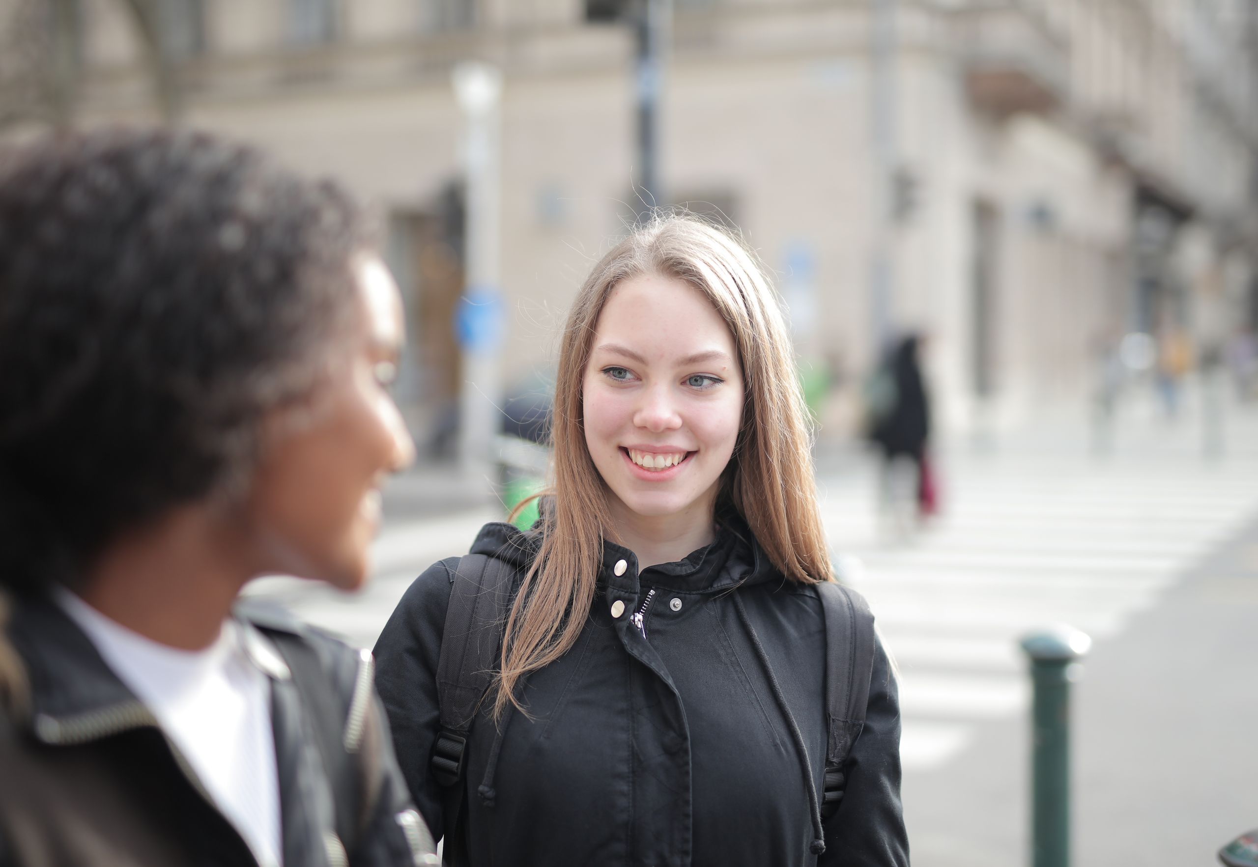 Two young women smiling and talking on a city street during the day.