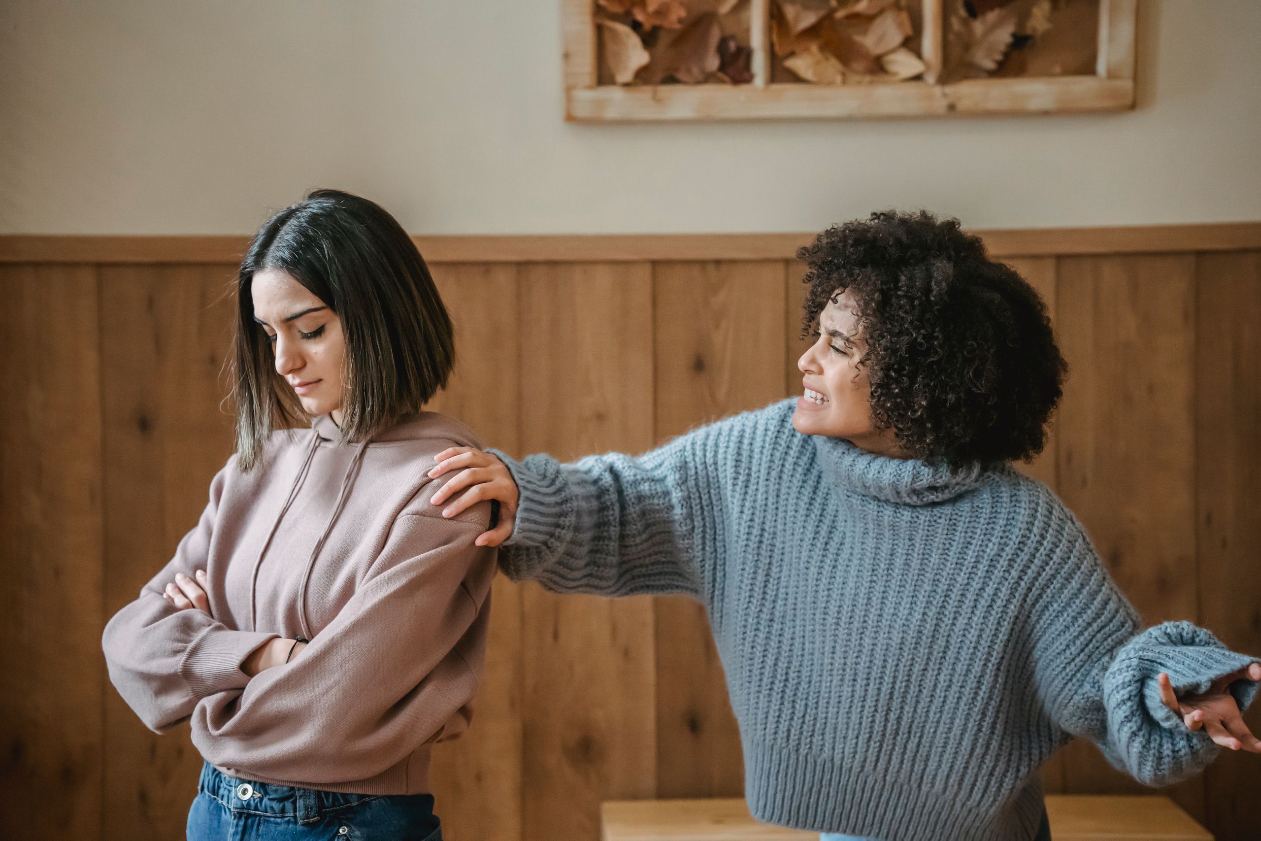 Unhappy young multiethnic female friends in casual outfit arguing with hand on shoulder while standing in bright house near wall
