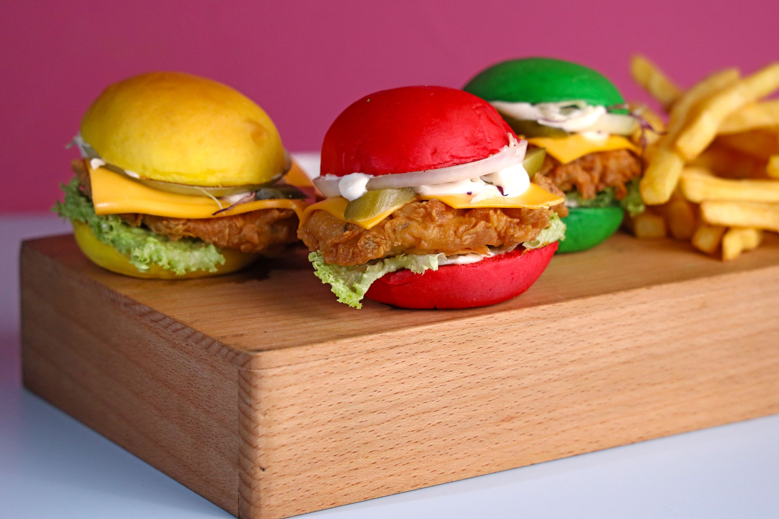 Vibrant chicken sandwiches with multicolored buns and fries on a wooden board.