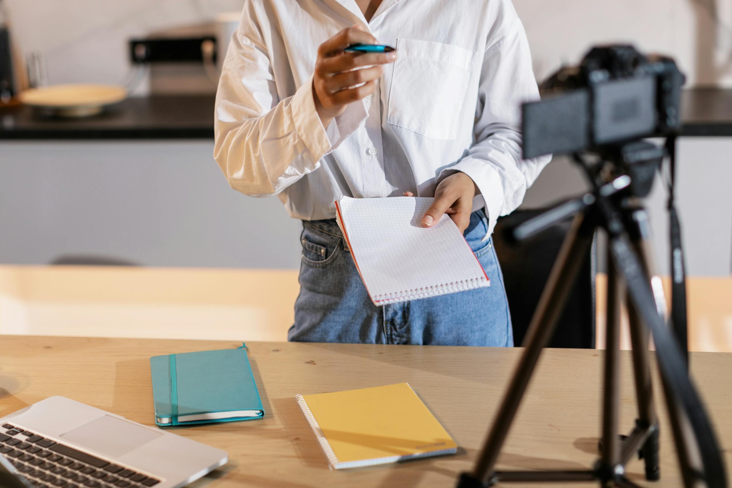 Vlogger recording content with a camera, holding notes and using a laptop in a modern setting.