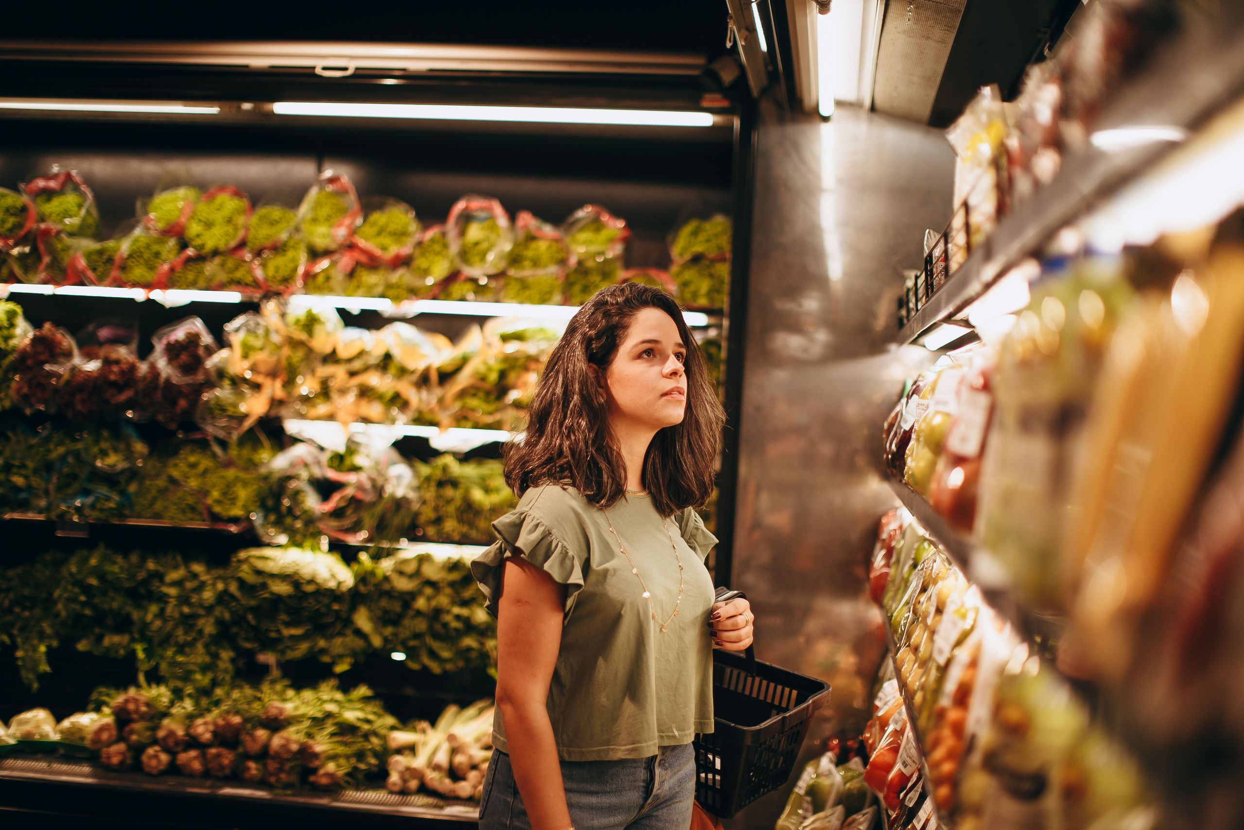 Woman browsing fresh produce with basket in supermarket. Indoor shopping scene.