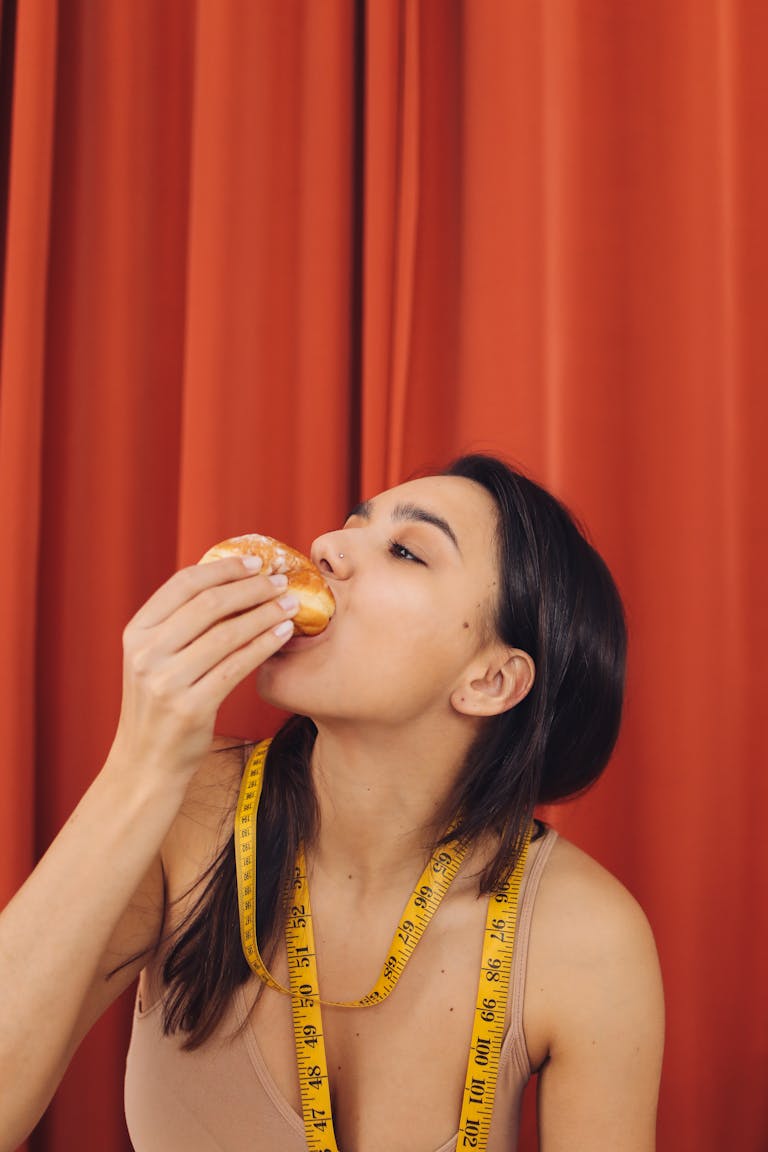 Woman eating pastry with measuring tape around neck in front of red curtain.
