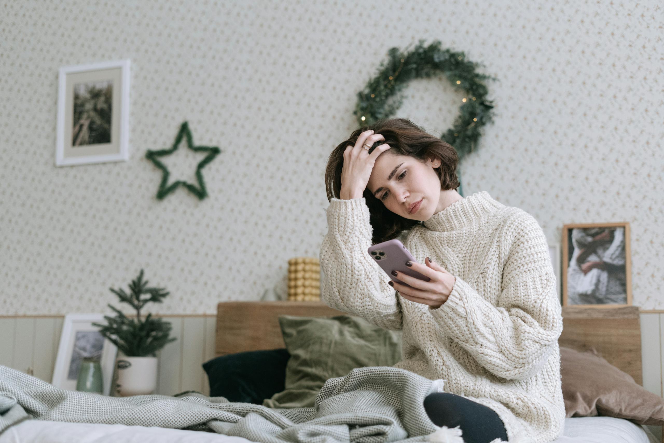 A woman in a knitted sweater is using smartphone while relaxing in a cozy bedroom.