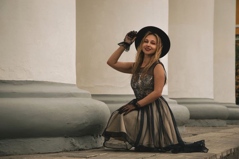 Woman in vintage dress and hat kneels outdoors, exuding timeless elegance.