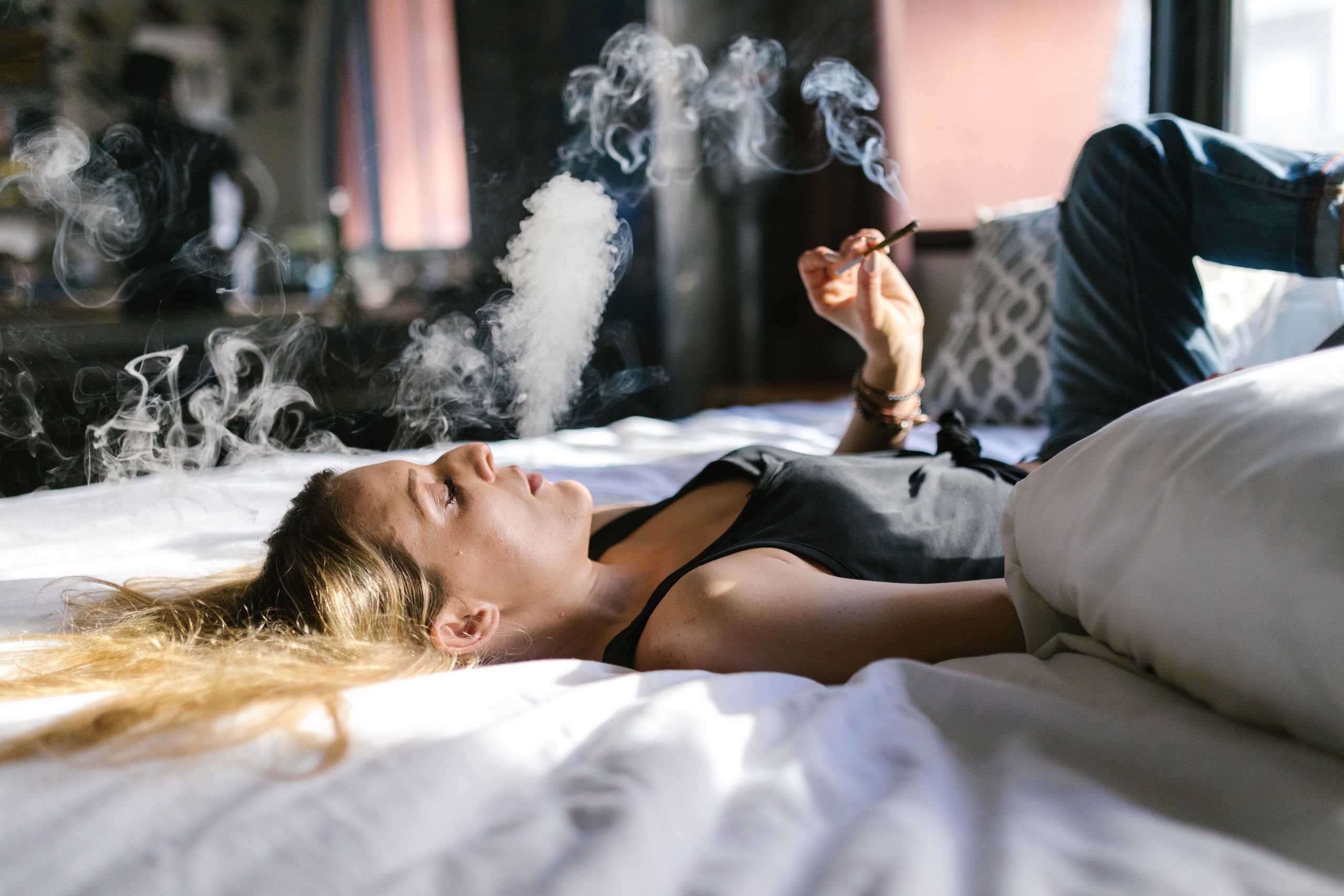 Woman lying on a bed smoking cannabis indoors, with smoke creating a serene atmosphere.