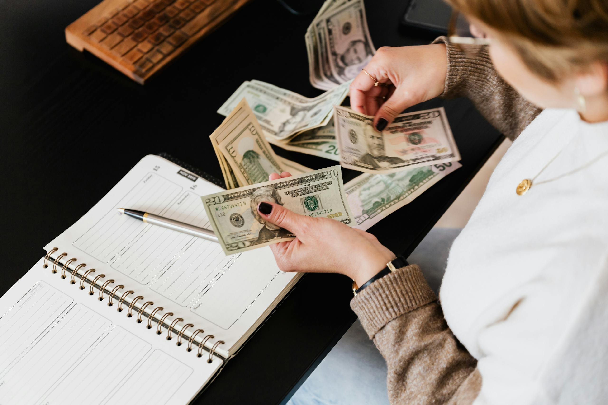 Woman organizing finances, counting dollar bills at desk with open notebook.