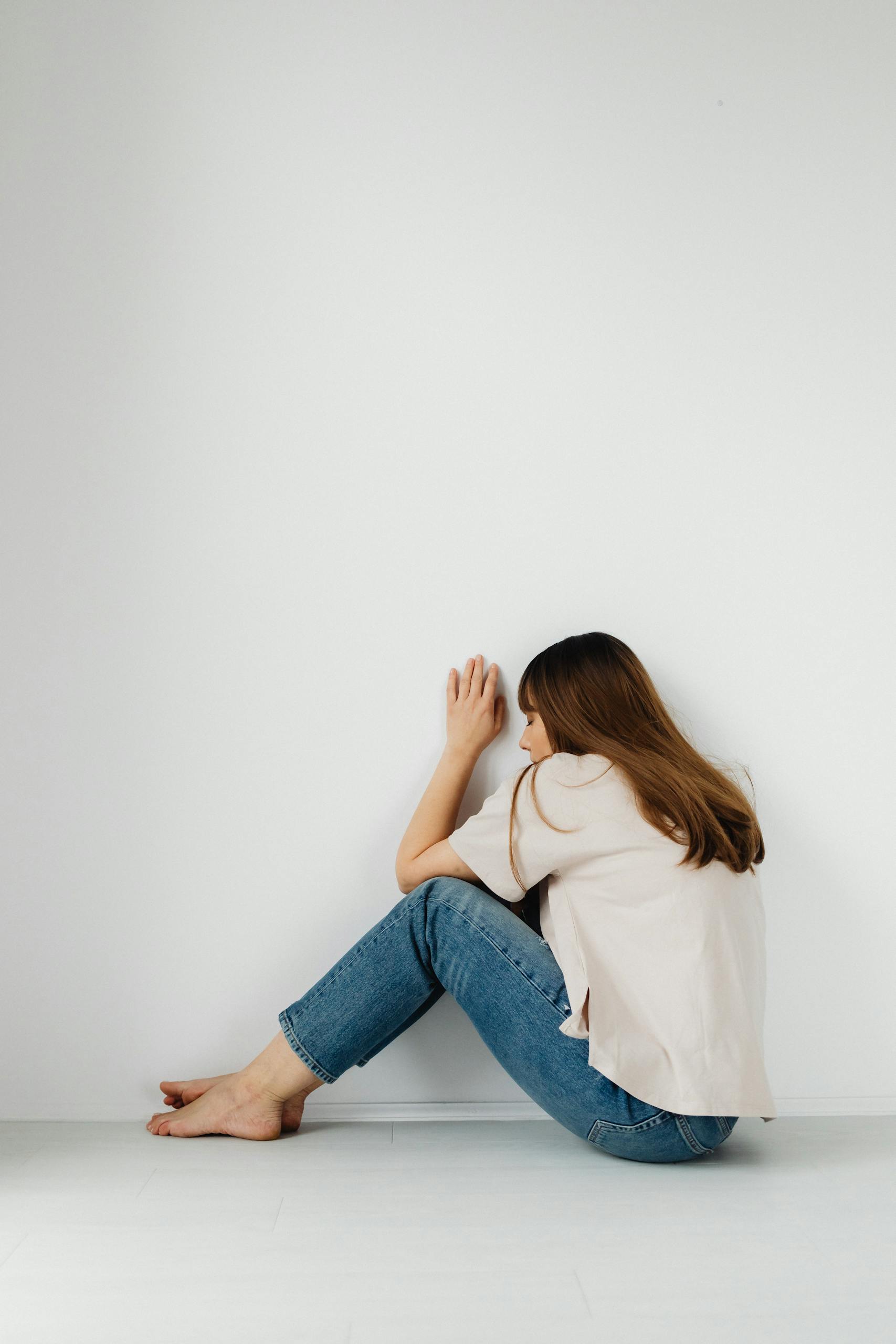 Woman sitting barefoot on floor in simple white room, deep in thought.