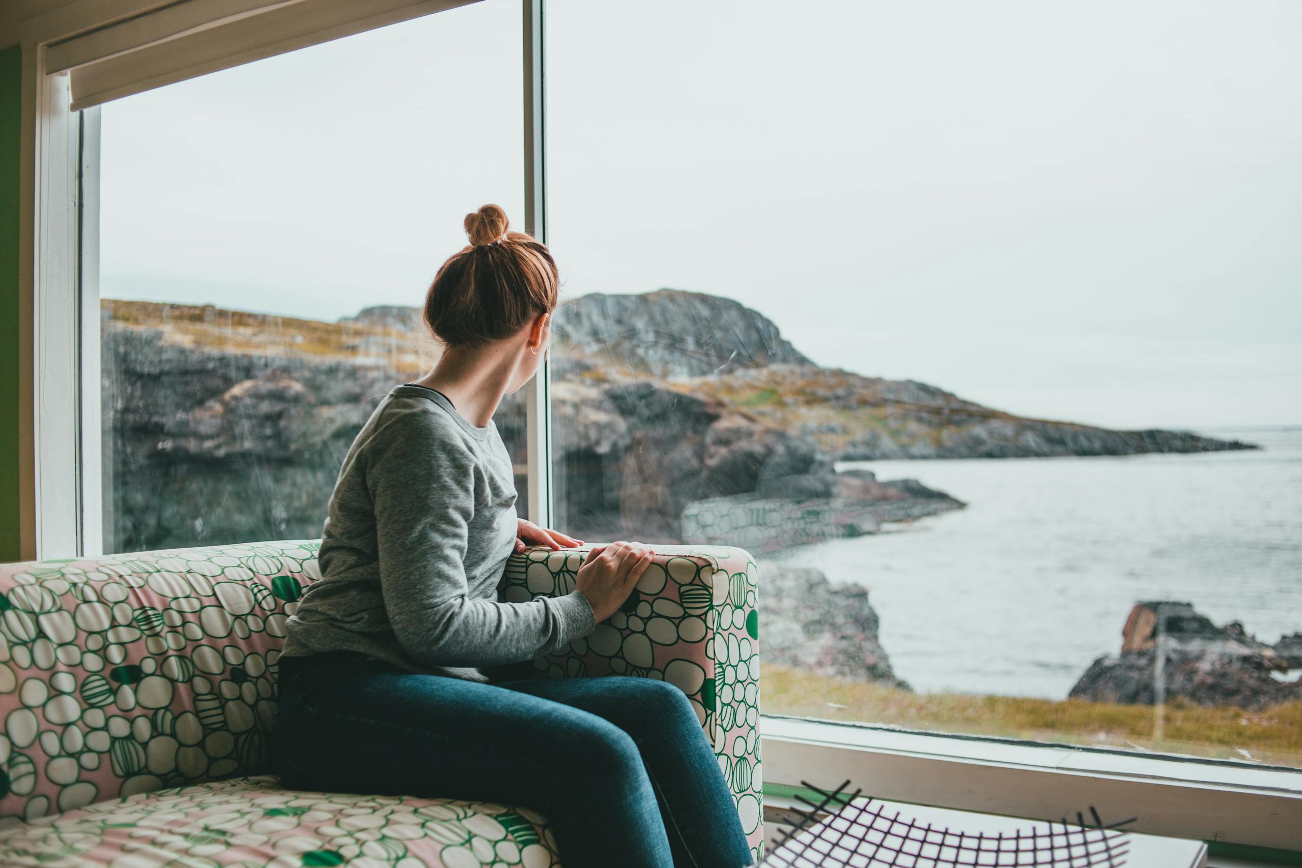 Woman sitting indoors gazing outside through a large window at a scenic rocky coastline.