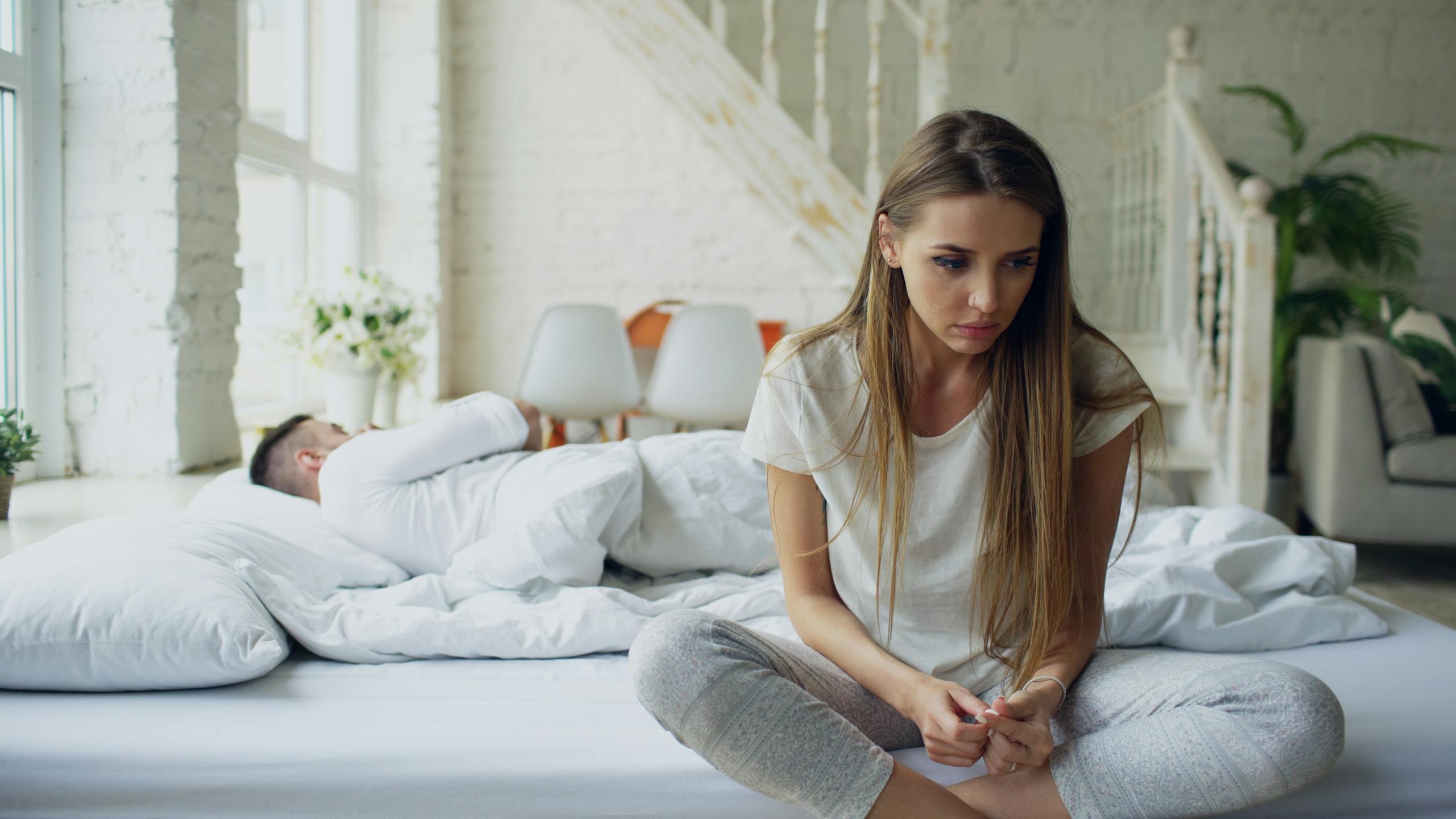 Woman sitting on bed looking thoughtful while man sleeps, symbolizing relationship tension.
