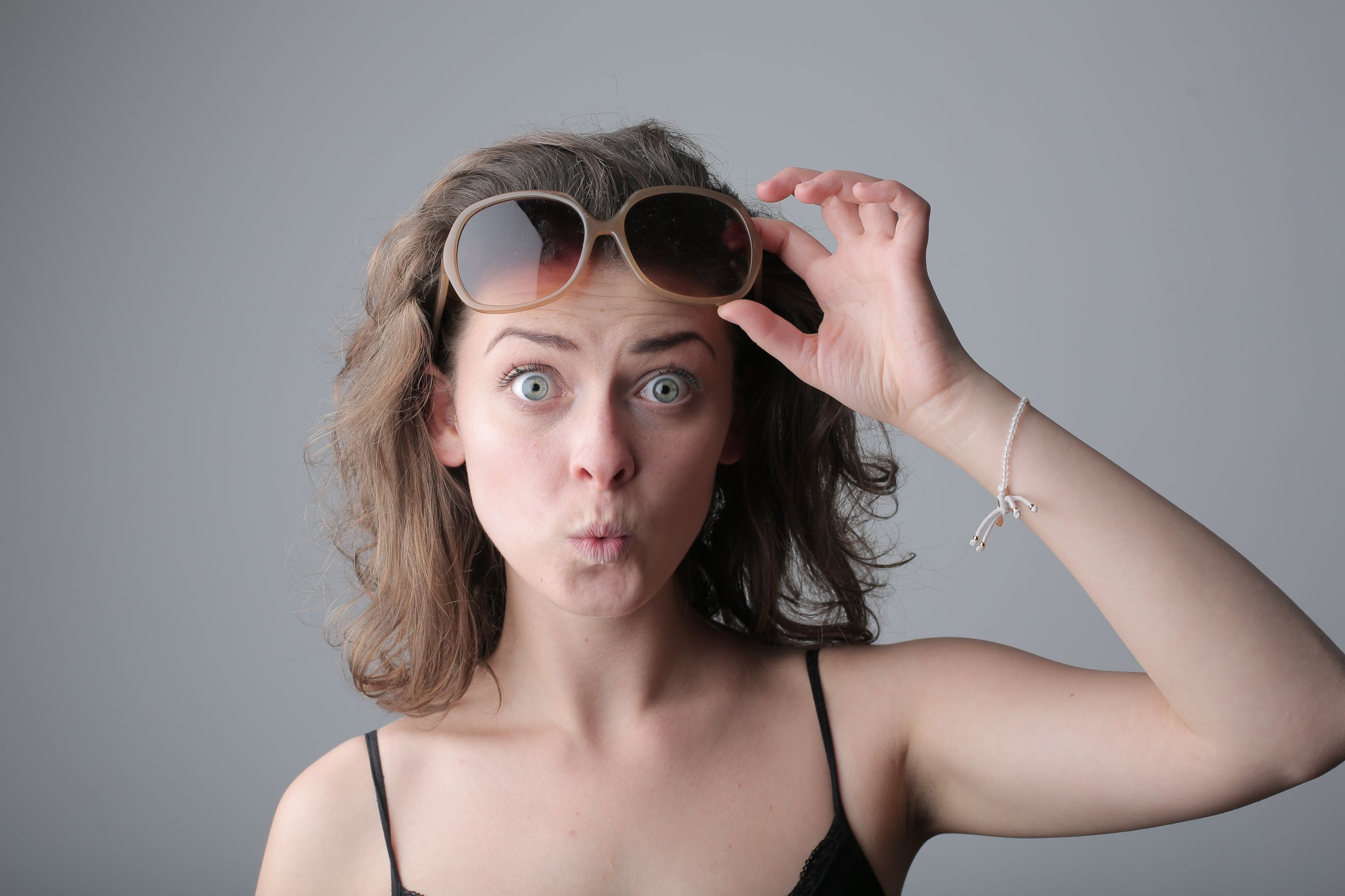 Woman with surprised expression lifting sunglasses indoors. Casual and playful mood.