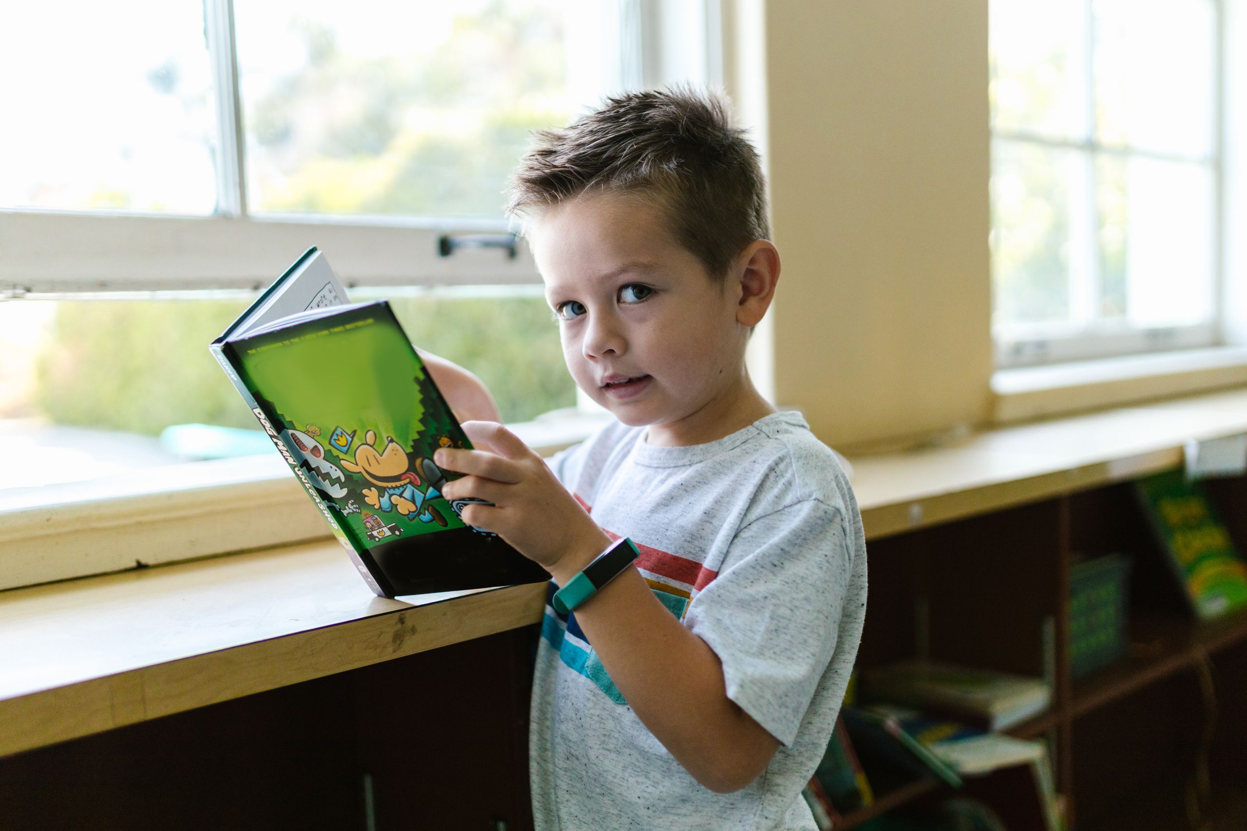 Young boy holding a colorful book while standing by a window, enjoying reading time.