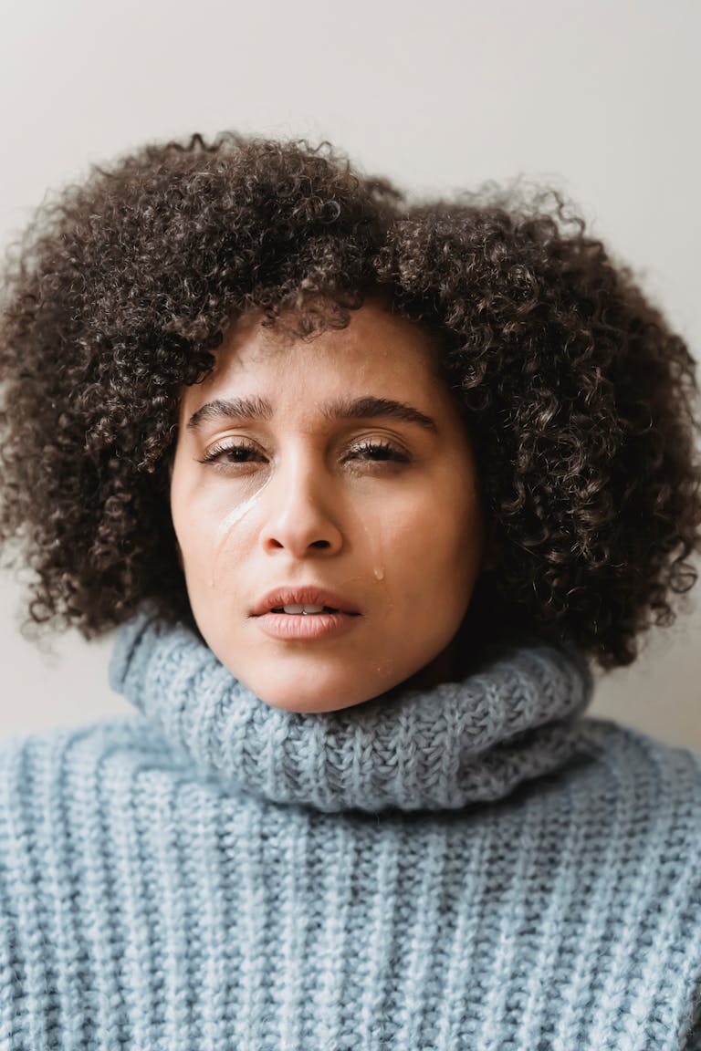 Young crying curly haired ethnic female with tears on cheek and opened mouth in warm pullover looking at camera against white background