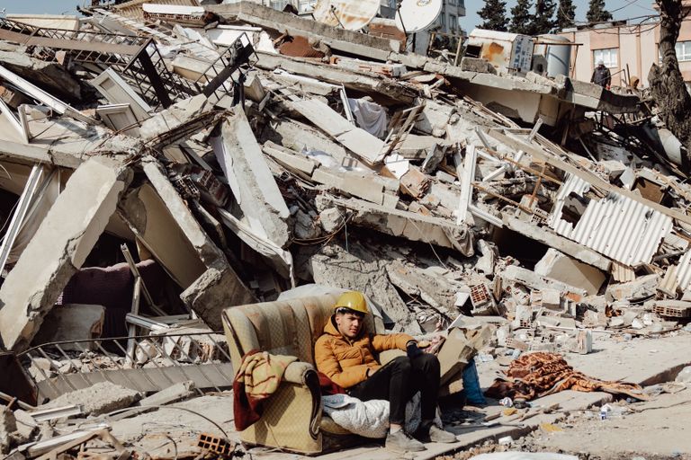 Young man in helmet on a sofa surrounded by destroyed buildings after earthquake.