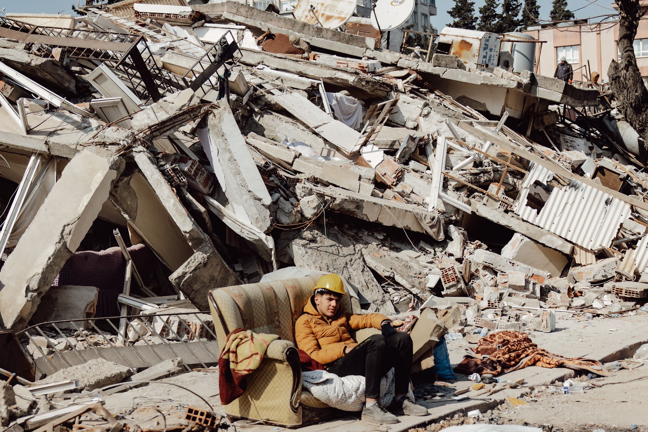 Young man in helmet on a sofa surrounded by destroyed buildings after earthquake.