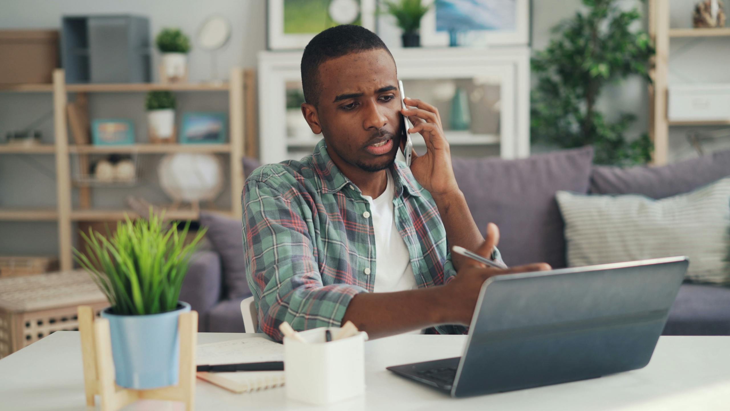 Young man multitasking with phone and laptop in a cozy home office environment.