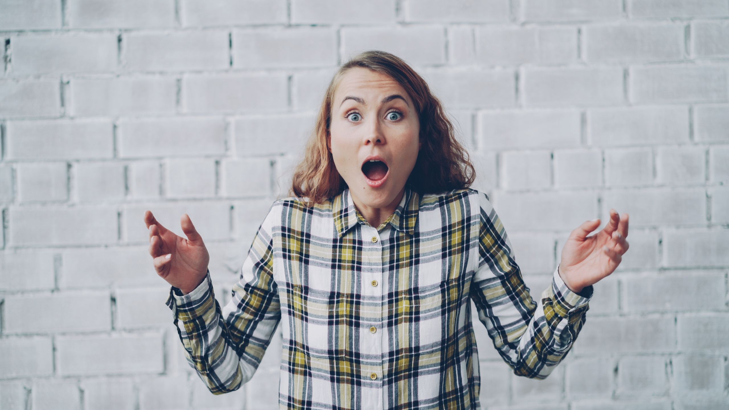 Young woman in plaid shirt expressing surprise against brick wall backdrop.