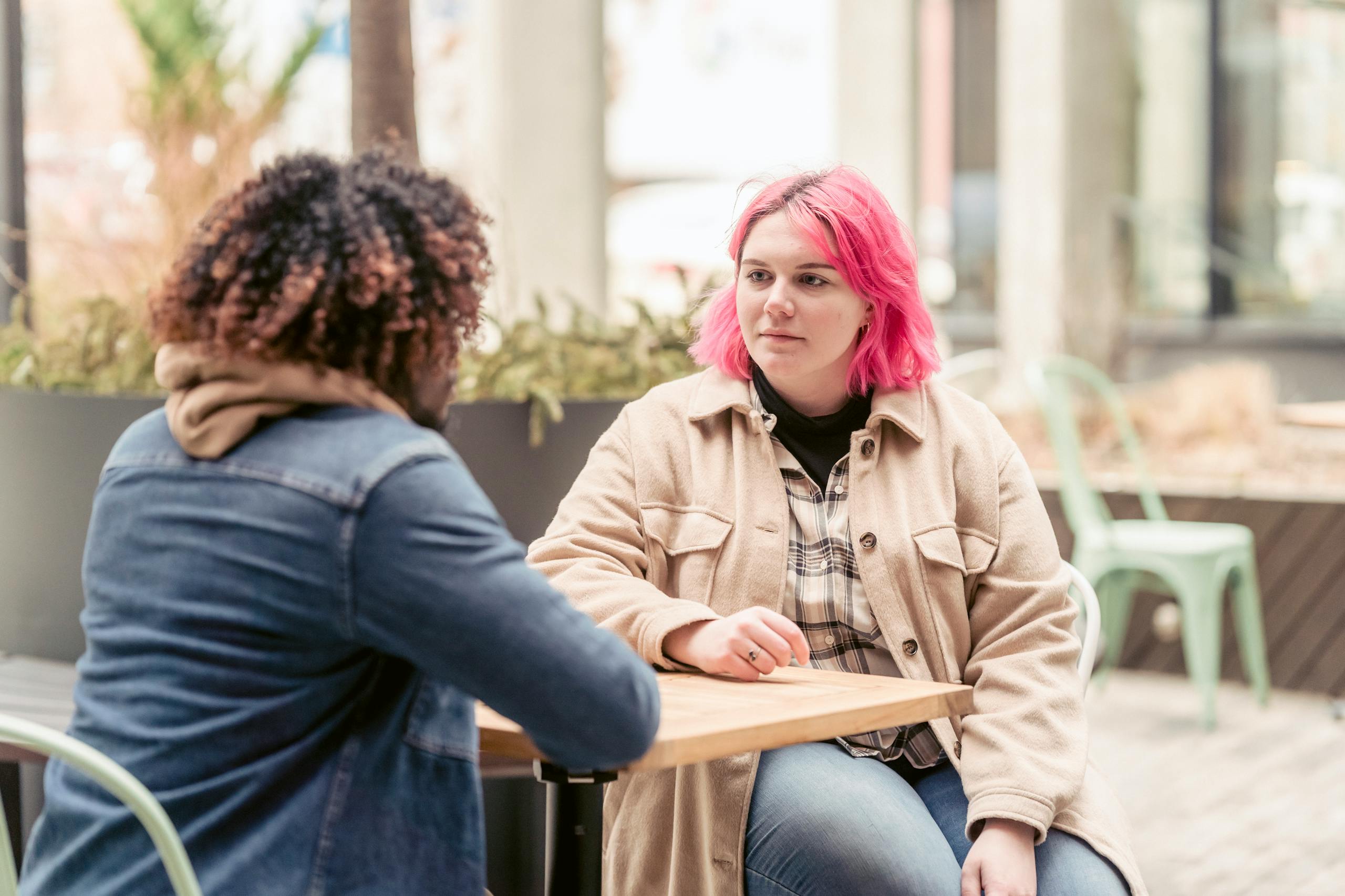 Young woman with pink hair listening to black man while sitting at table in weekend in outdoor cafeteria