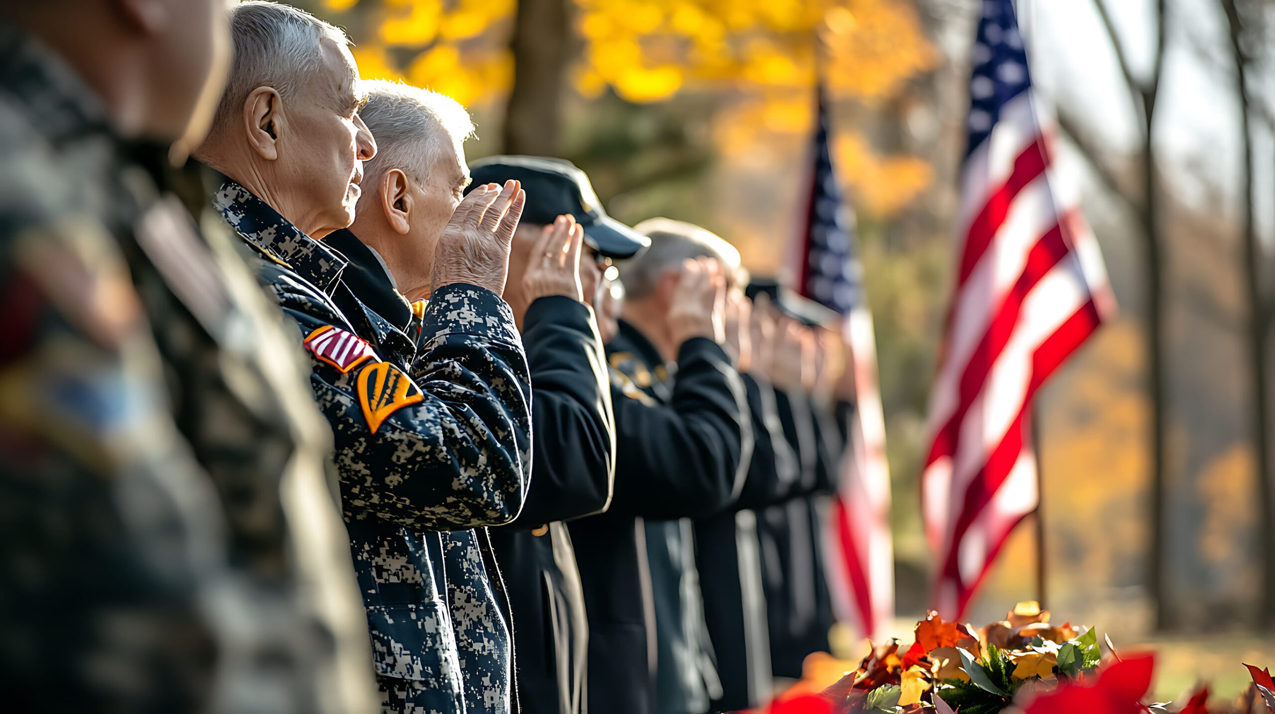 A group of soldiers in military uniform salutes, standing in a line with an American flag in the background, during a ceremony.