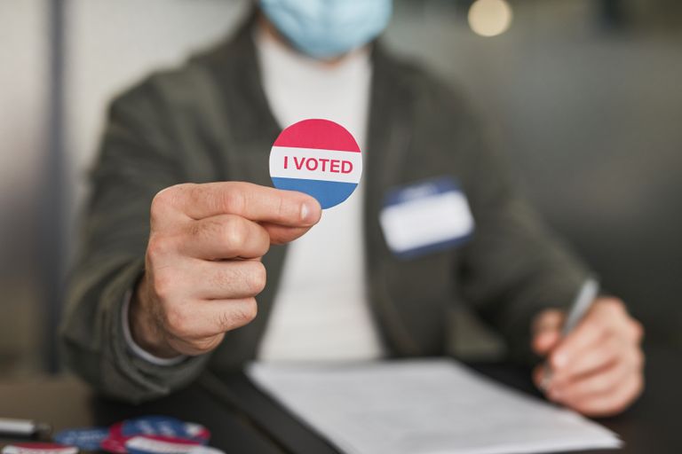 A person in a mask holding an 'I Voted' sticker, emphasizing civic engagement.