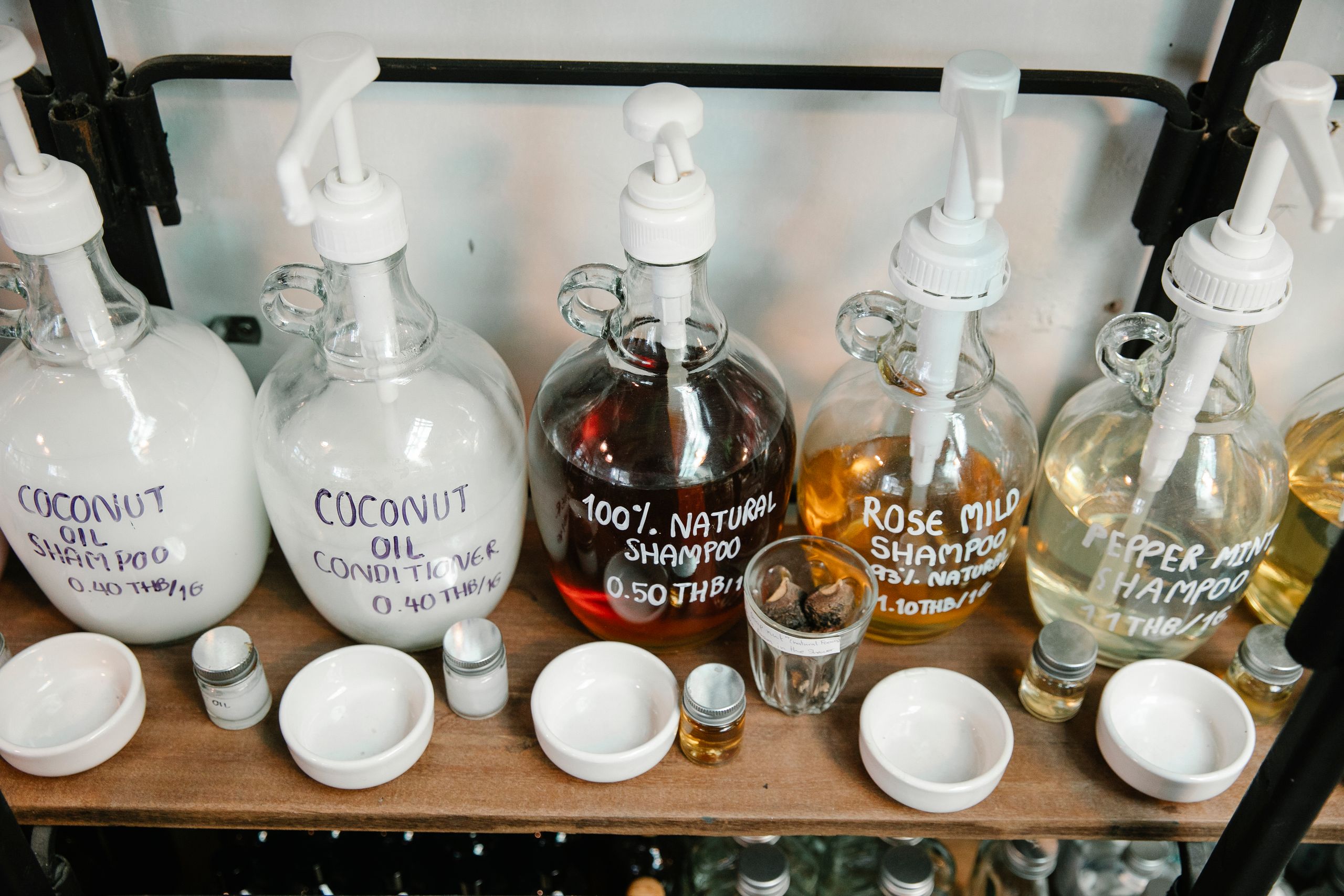 Collection of big dispensers of various multicolored shampoo placed near glass jars and white bowls on wooden shelf