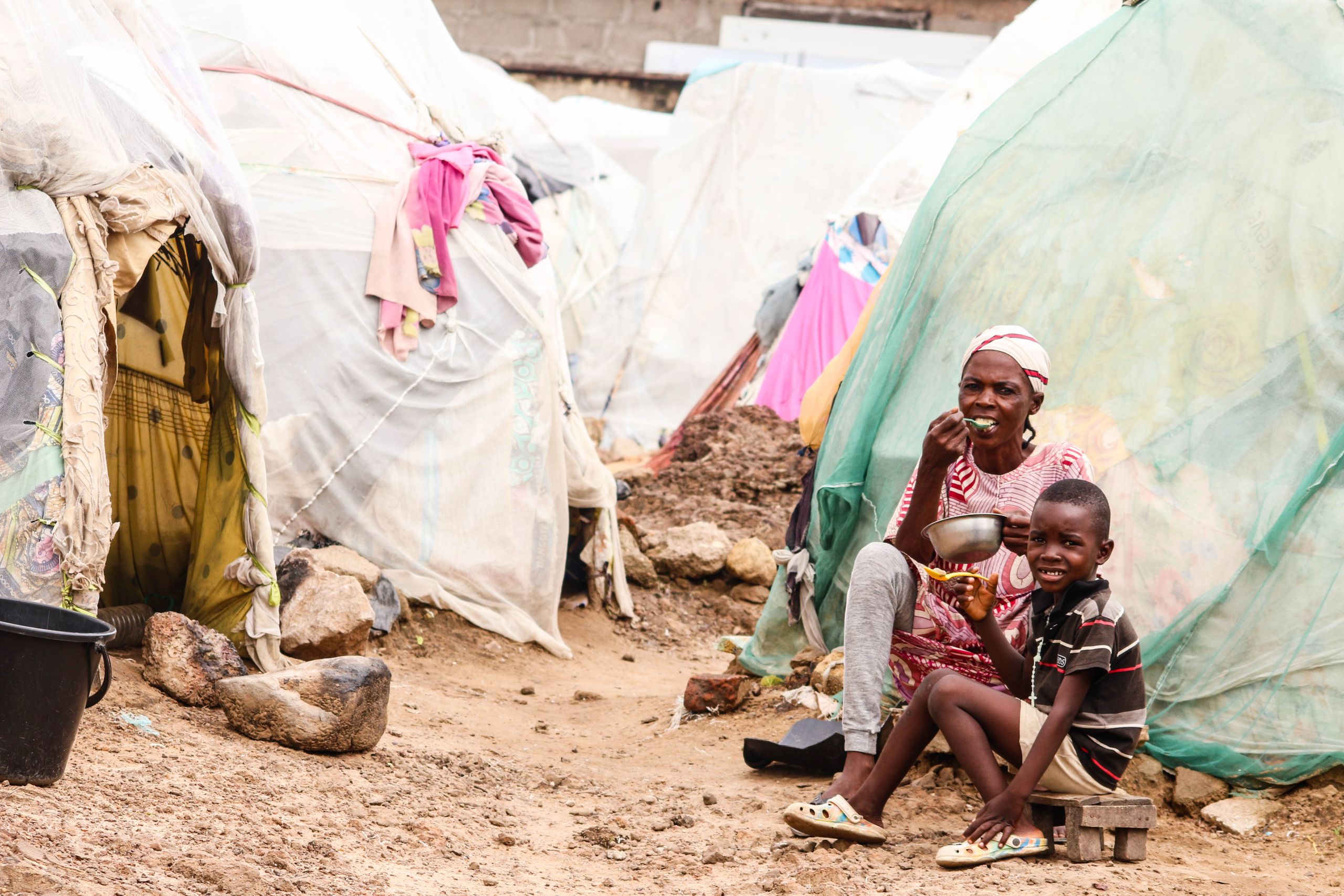 Mother and son enjoying a meal in a rustic village scene with makeshift housing.