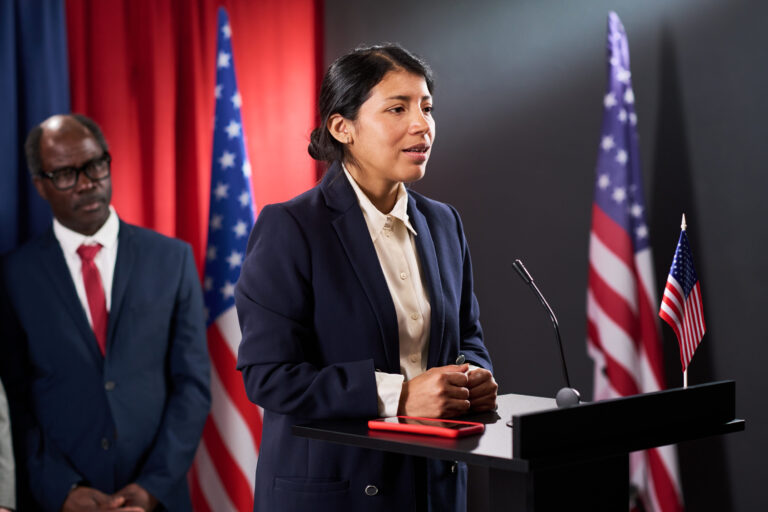 Medium shot of Latin young politician speaking at podium during campaign event, addressing public policy and leadership. Ideal for election marketing, civic engagement