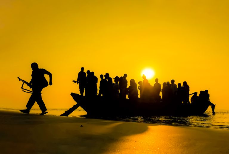 Dramatic silhouette of a crowded boat and individuals against a golden sunset on the water's edge.