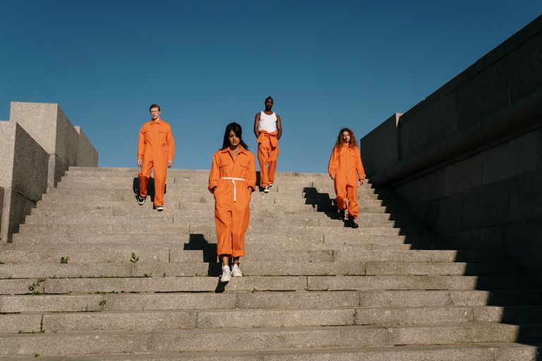 Four diverse individuals in orange jumpsuits descending outdoor stairs under bright blue sky.