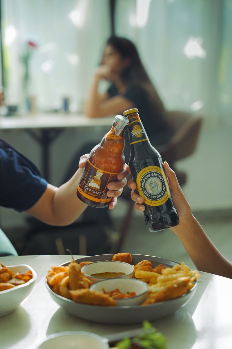 Two people toasting with craft beer bottles over a table of snacks.