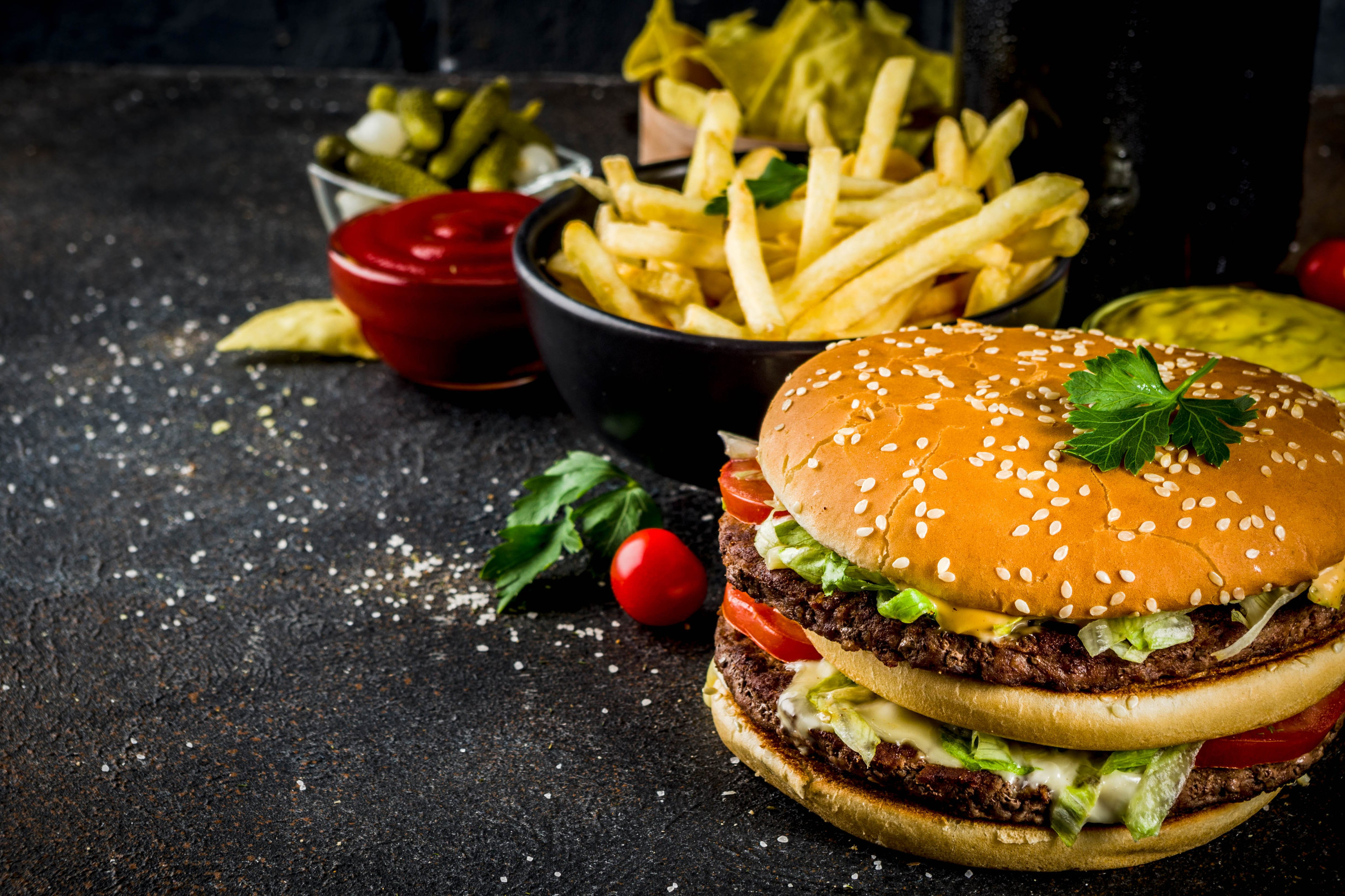 Various party food, Hamburgers, French fries, potato chips, pickled cucumbers, onions, tomatoes and cold beer bottles, rusty black concrete background copy space