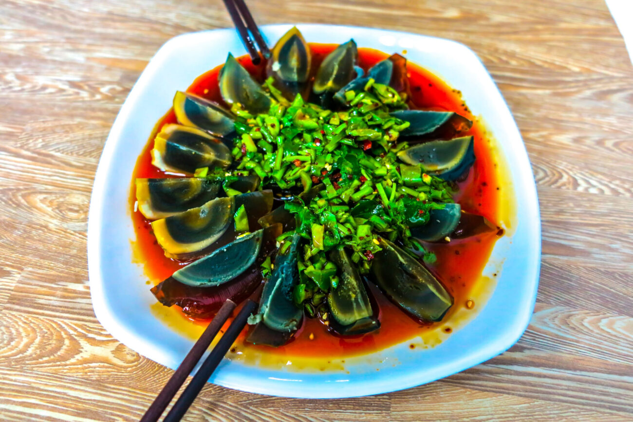 A vibrant platter of century eggs garnished with fresh green onions, all resting in a rich, spicy sauce, inviting food lovers to explore a unique culinary experience. Image Credit: alexela82/ 123RF