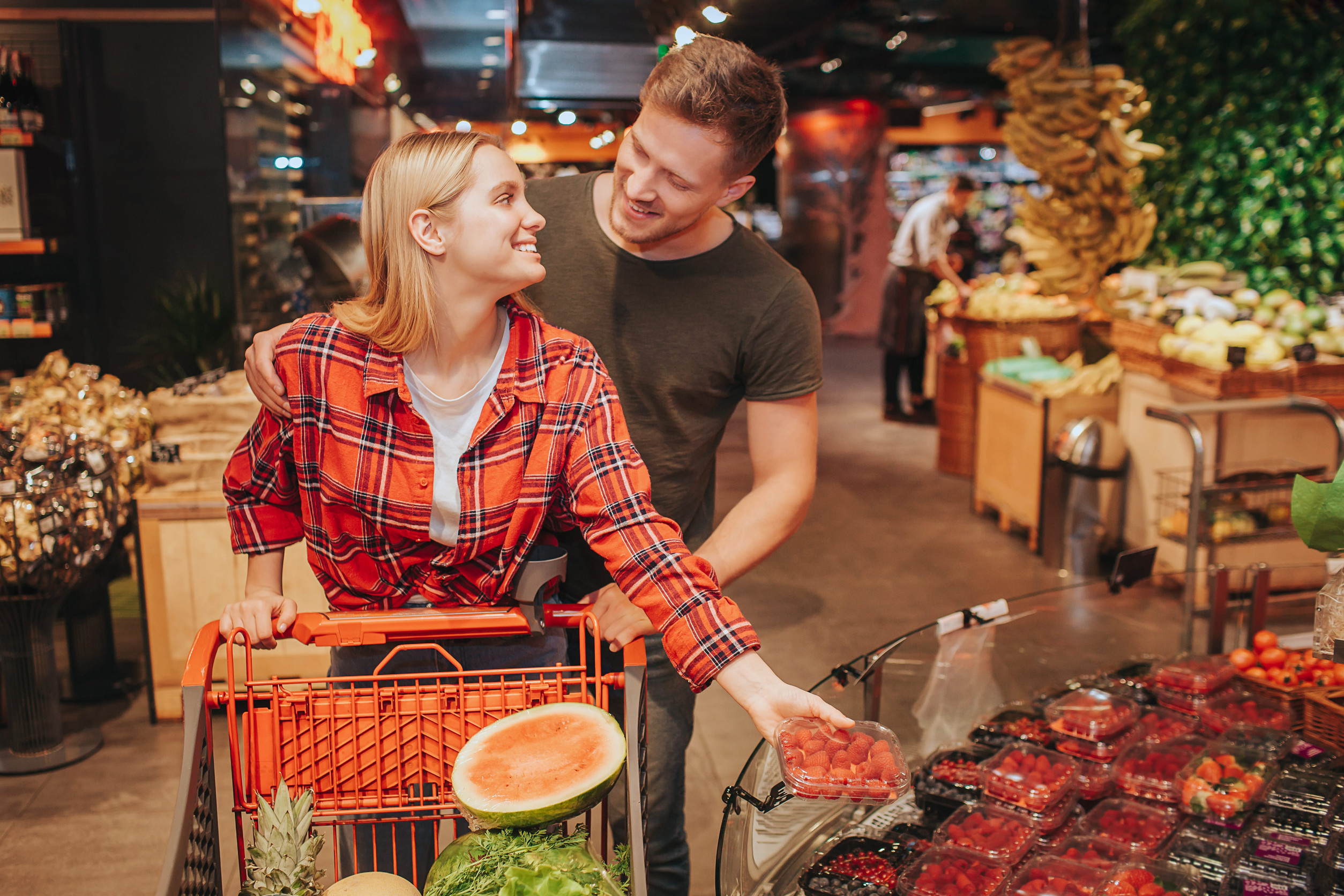 Young couple in grocery store. She get plate with berries and look at man. Guy Stand behind her and hold trolley with hand. Smile to each other.