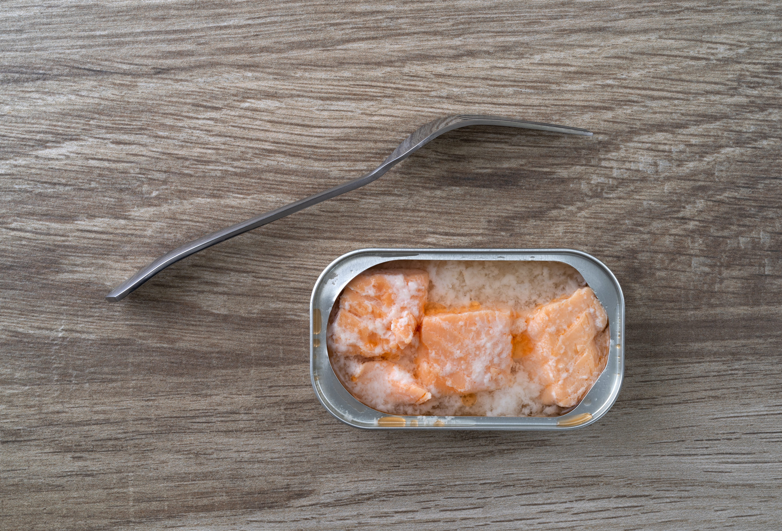 Open tin of salmon in water with a fork to the side on a countertop illuminated with natural lighting.