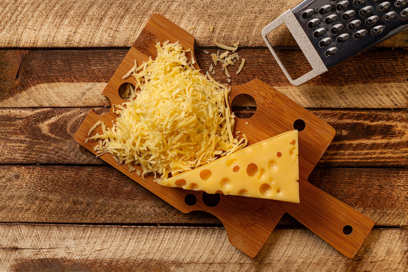 A delightful scene showcasing freshly grated cheese alongside a wedge of creamy, yellow cheese on a rustic wooden board, perfect for culinary inspiration. Image Credit: amashukeli/ 123RF