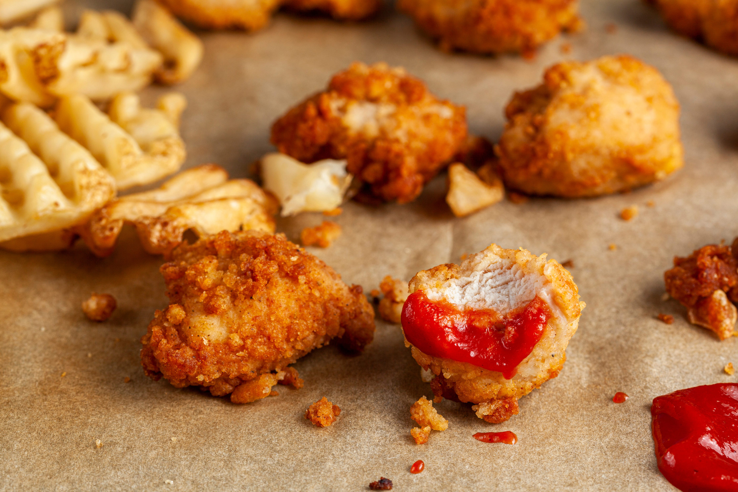 Quick convenient fast food image with close up selective focus of bitten deep fried chicken nuggets dipped into red tomato ketchup on baking paper. Ruffled waffle potato fries are in the background.