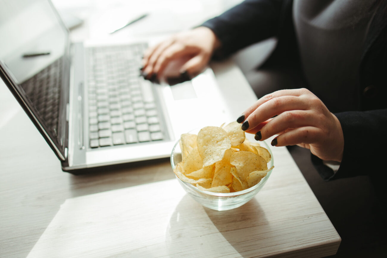 Amidst the hustle of work, a moment of indulgence is found in a simple bowl of chips. As fingers reach for the crispy snack, the laptop hums with productivity, blending leisure and professionalism in a harmonious workspace. Image Credit: jetsam86/ 123RF