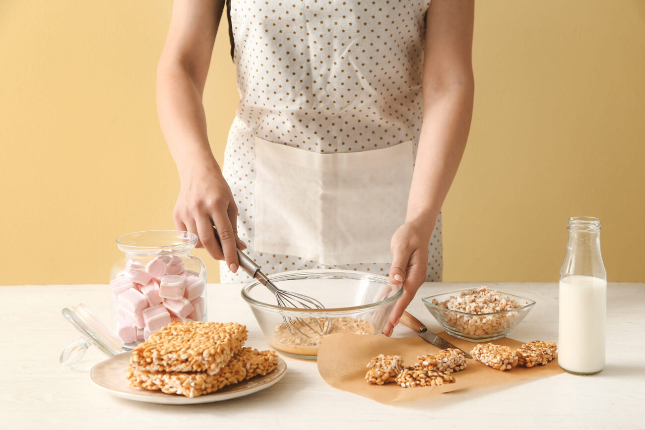 Woman cooking crispy rice bars at table, blending ingredients with care while a jar of marshmallows and a bottle of milk sit nearby. Image Credit: serezniy/ 123RF