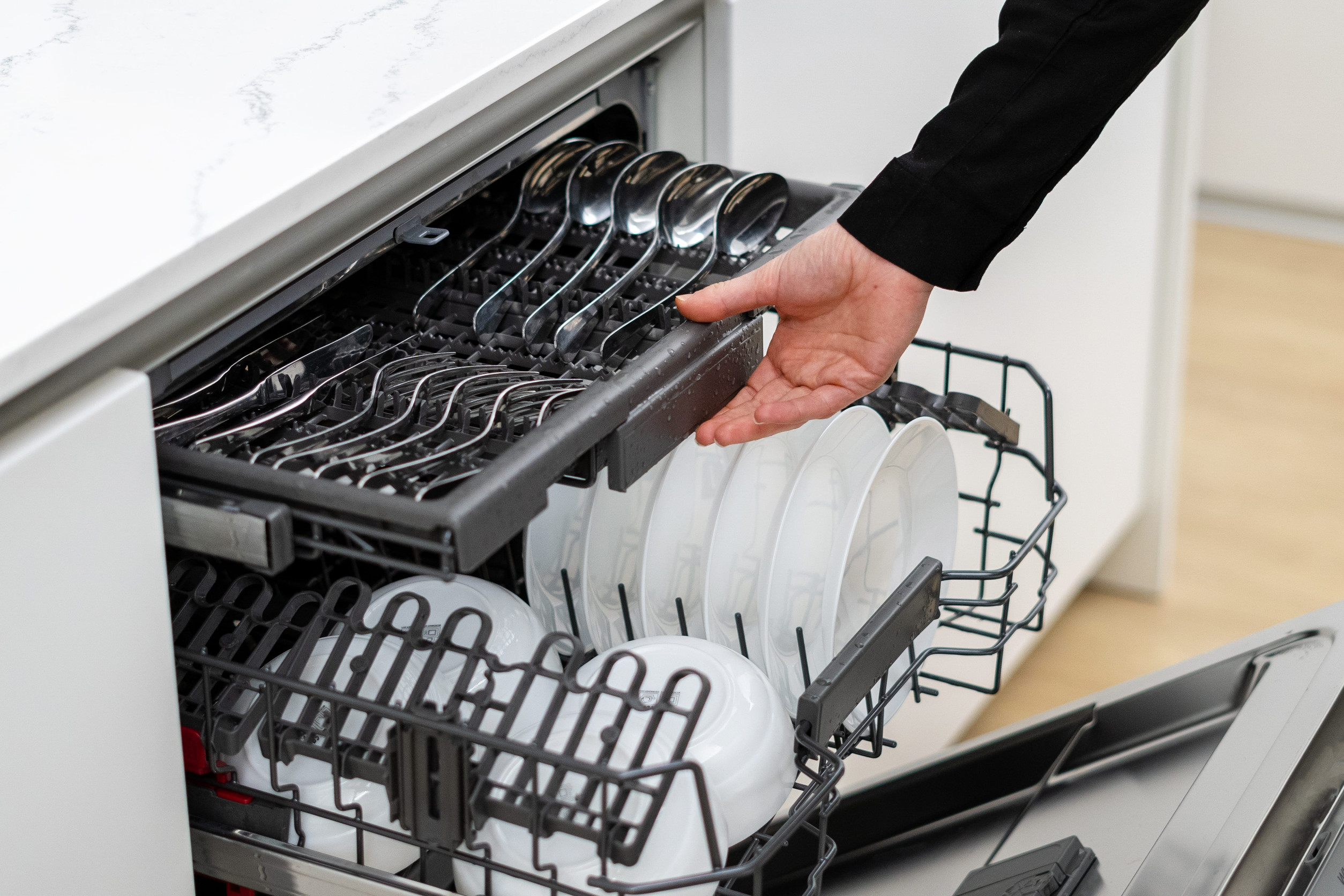Built in dishwasher with open door, cropped shot of woman loading it with dishes and tableware, pushes drawer full of spoons and forks inside. Kitchen furniture with built in electric appliances