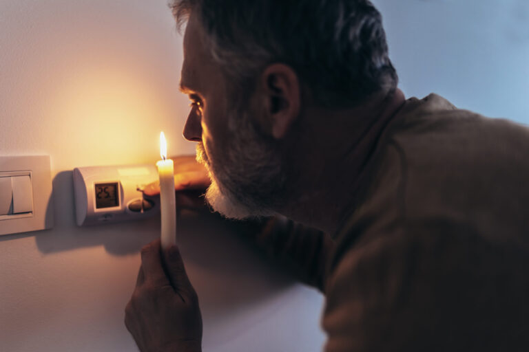 Energy crisis. Man in complete darkness holding a candle to investigate thermostat during a power outage. Blackout concept.