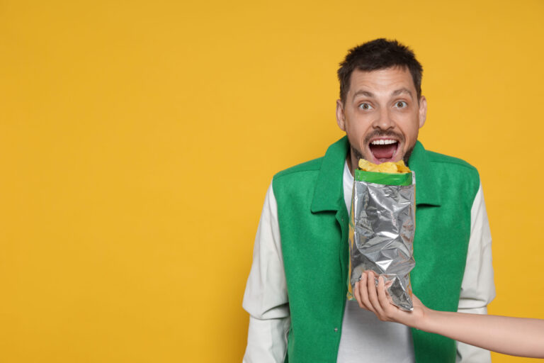 Woman offering tasty potato chips to excited man on orange background, space for text