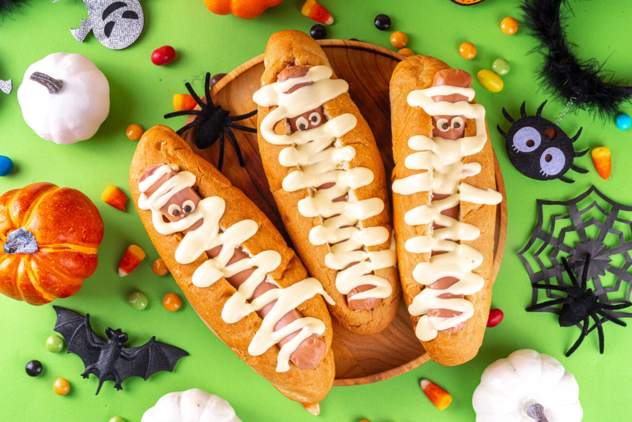 A spooky delight for Halloween, these mummy hot dogs, wrapped in creamy icing bandages, sit surrounded by festive treats and decorations, making for a fun and tasty celebration. Image Credit: bondd/ 123RF
