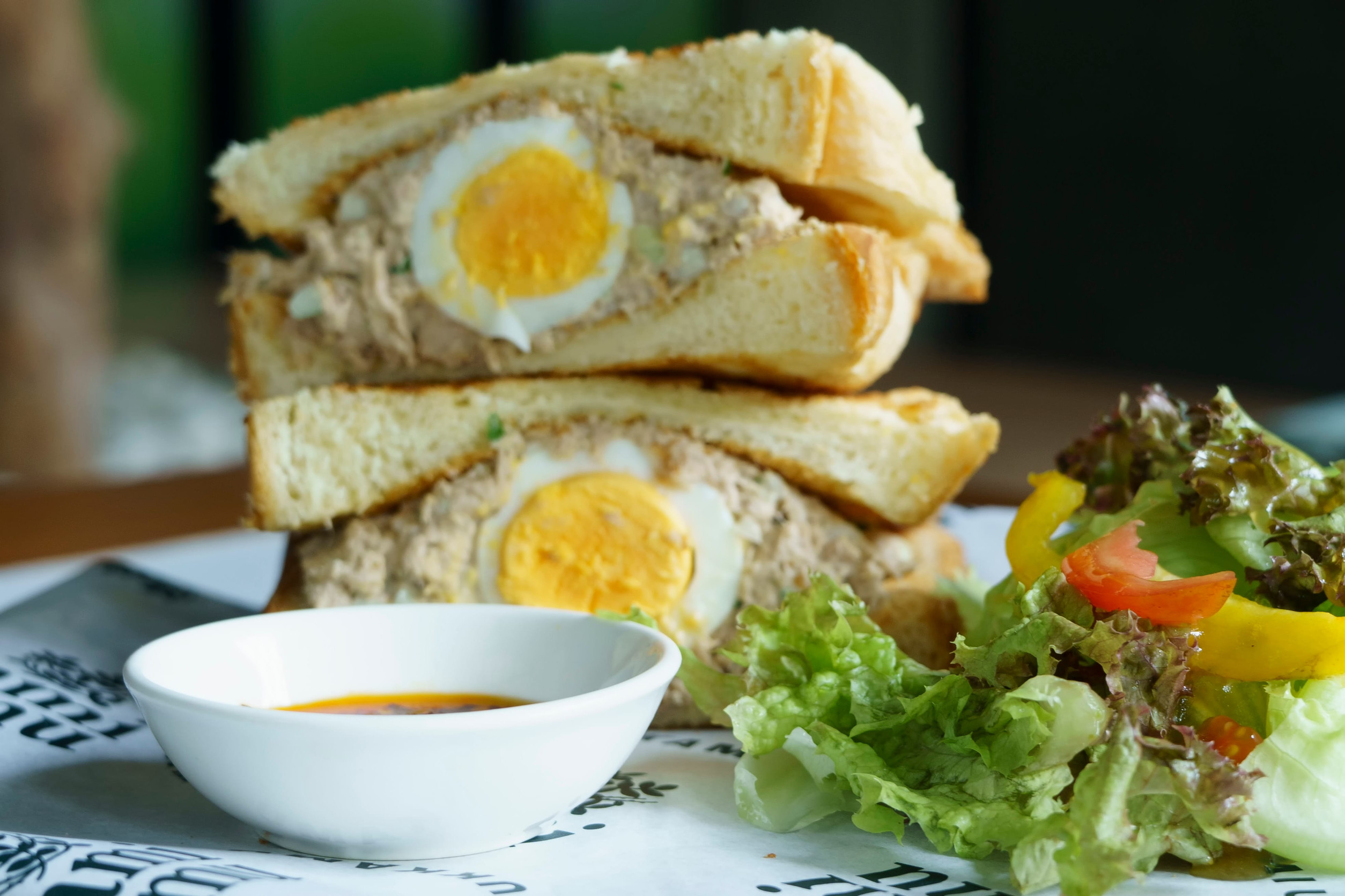 Crispy toasted bread filled with tuna salad and a halved hard-boiled egg, served with a side salad and a small bowl of dipping sauce.
