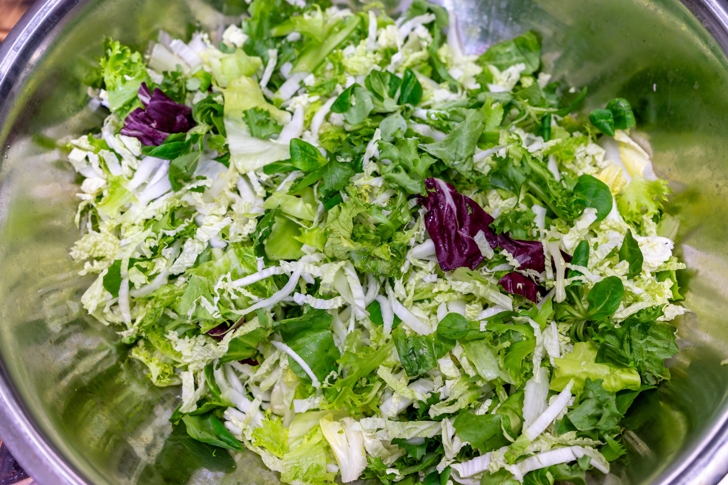 A large stainless steel bowl holds an assortment of freshly chopped green leaves, including romaine, lettuce, and herbs. The vibrant colors suggest a healthy meal option.