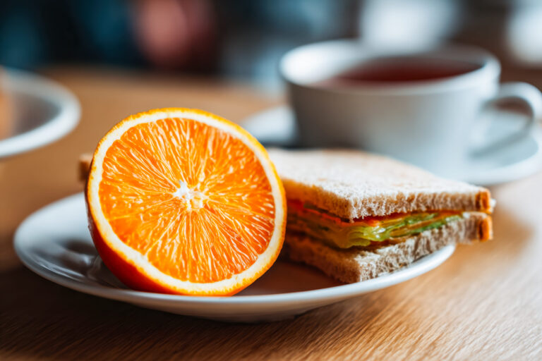 A vibrant orange wedge alongside a hearty sandwich and a warm cup of tea create a cozy, inviting scene perfect for breakfast or snack inspiration. Image Credit: mikkiorso/ 123RF