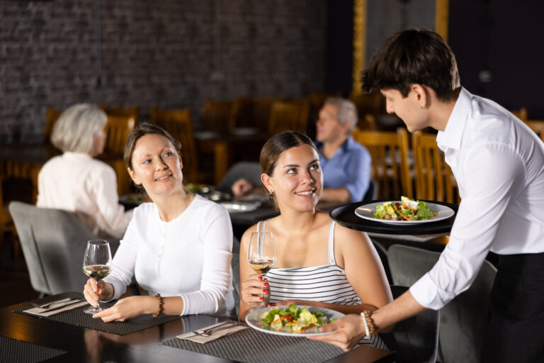 Young male waiter serving dish to two women in restaurant. Image Credit: jackf/ 123RF