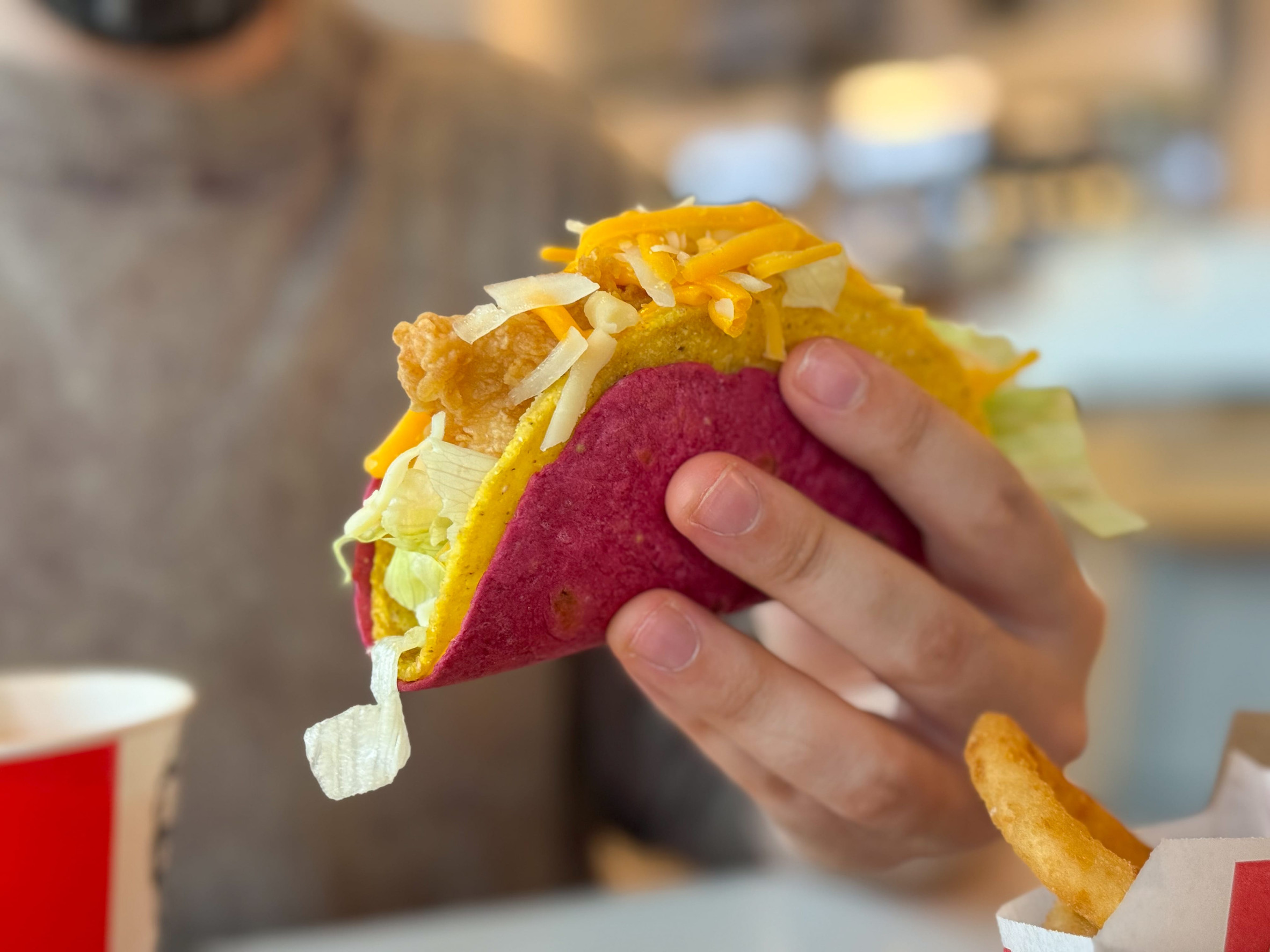 A person holds a colorful taco with cheese, lettuce and crispy filling inside a cafe. Fast food culture, casual dining, consumer lifestyle and modern gastronomy in urban environment.