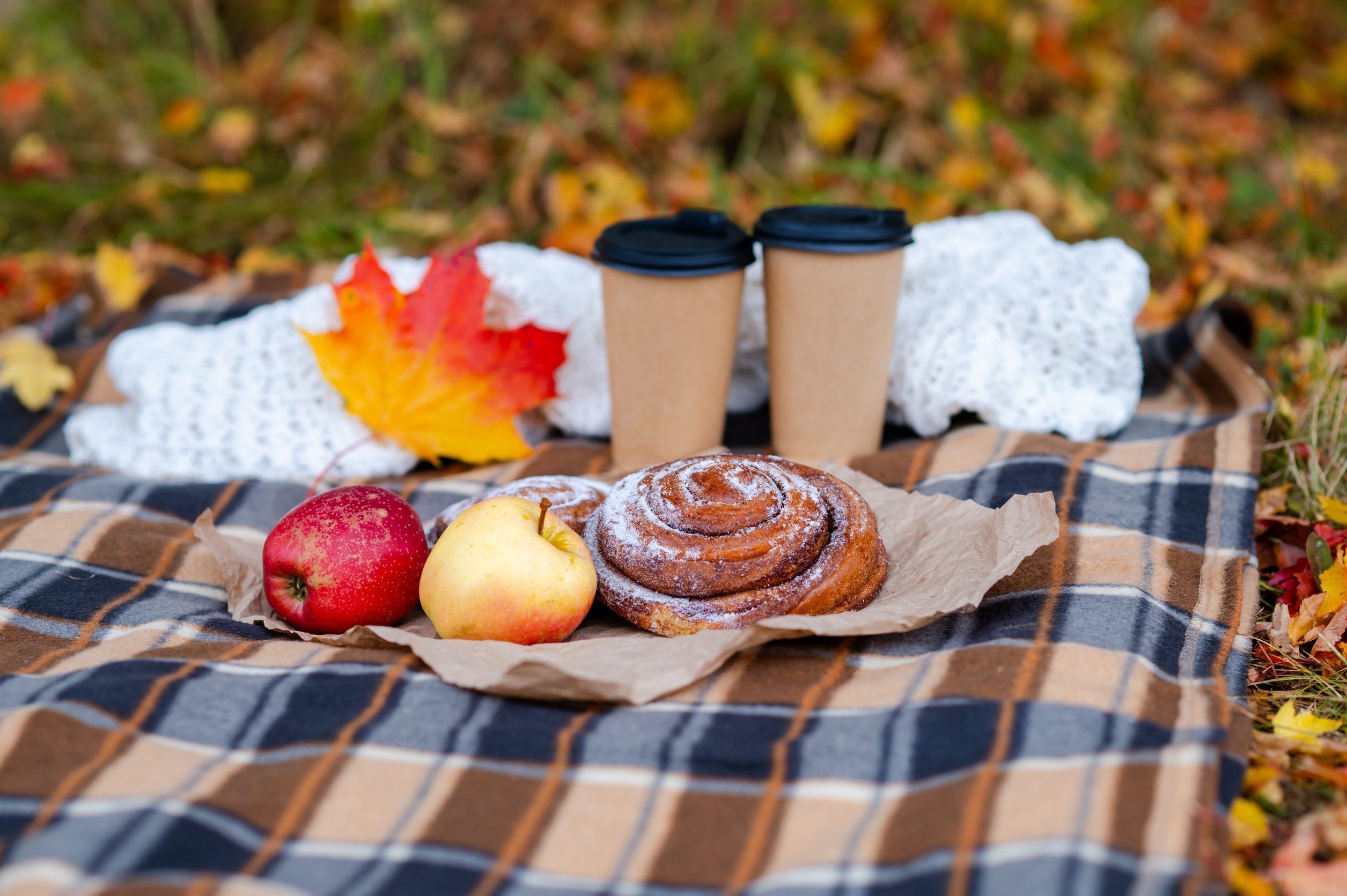 Autumn cozy picnic with a knitted sweater, plaid blanket, two paper cups of hot drinks, pastries, fresh apples, and colorful maple leaves falling around