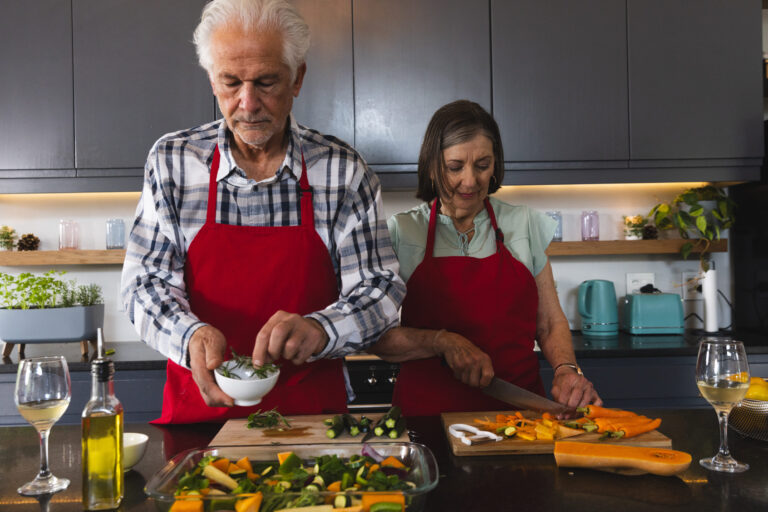 Senior couple cooking at kitchen island wearing red aprons, sprinkling herbs and slicing carrots. Cooking, kitchen, gourmet, culinary, preparation, rustic, wellness